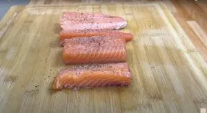 Four salmon fillets seasoned with black pepper in a cast iron skillet, ready for cooking on a wooden countertop.